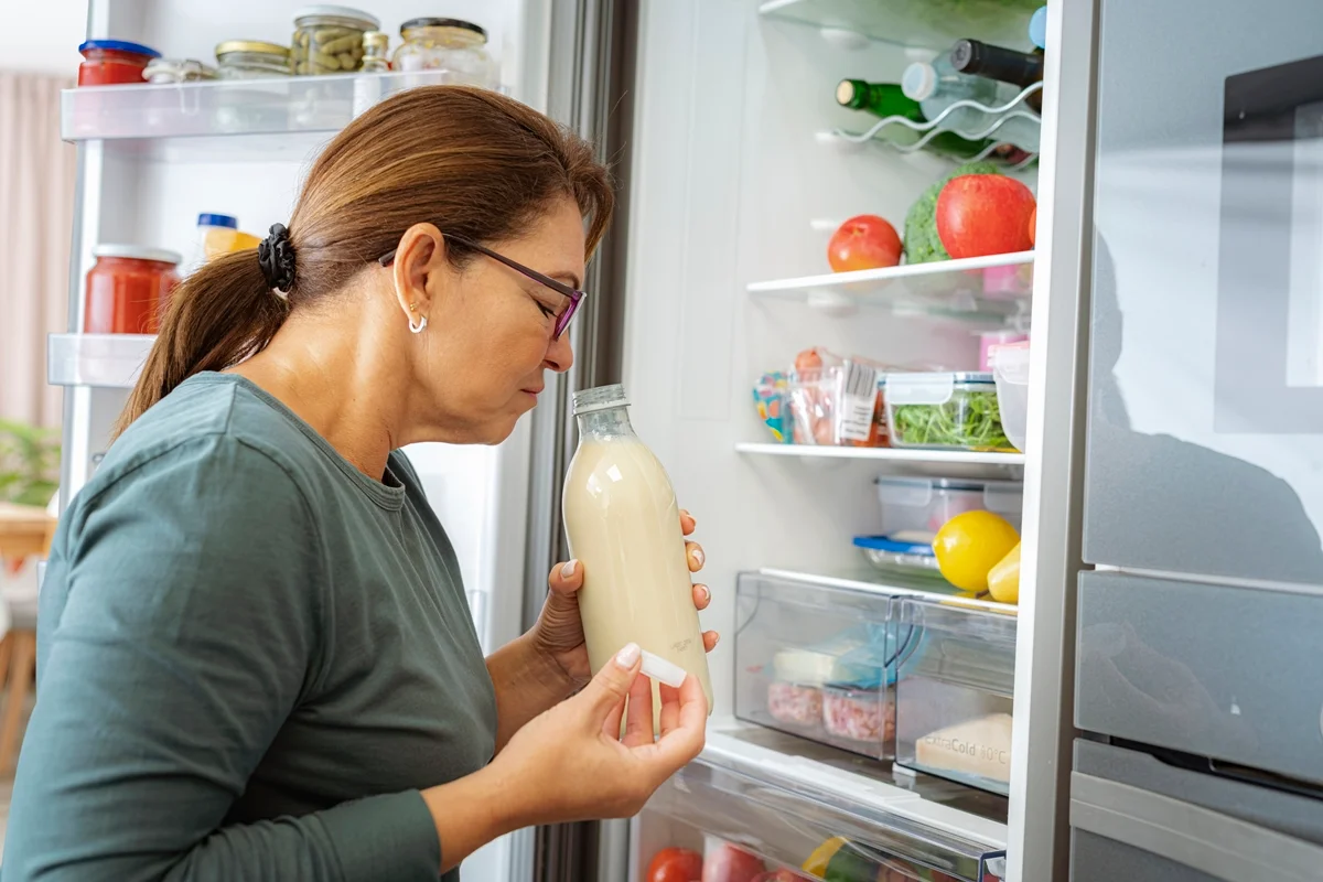 Foto colorida de mulher cheirando leite dentro de garrafa em frente a geladeira - Metrópoles